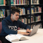 Indian student studying with a laptop and textbooks in a library while preparing to study abroad without IELTS